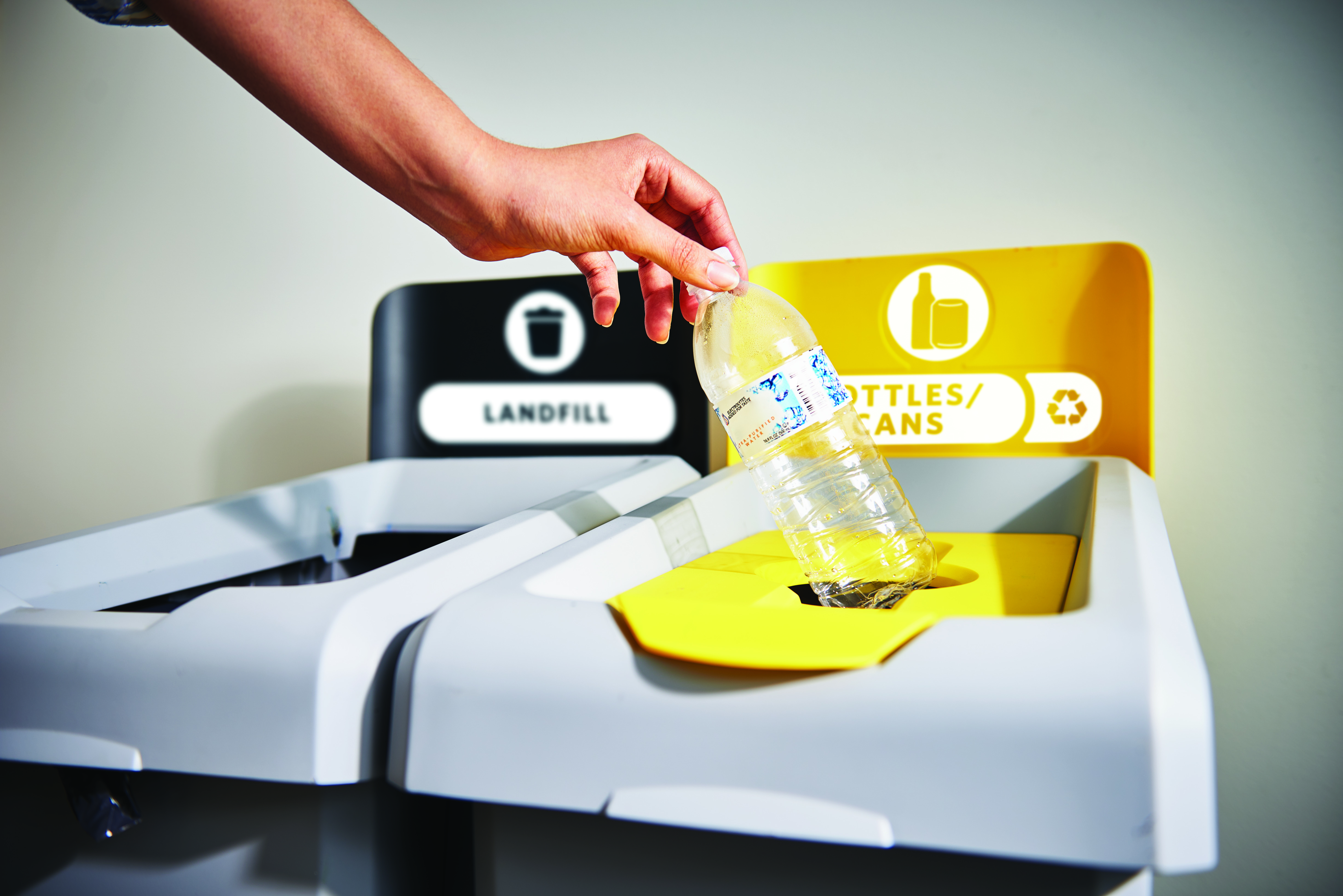 Hand placing plastic bottle into yellow recycling bin labeled ""Bottles/Cans"" next to a black landfill bin."
