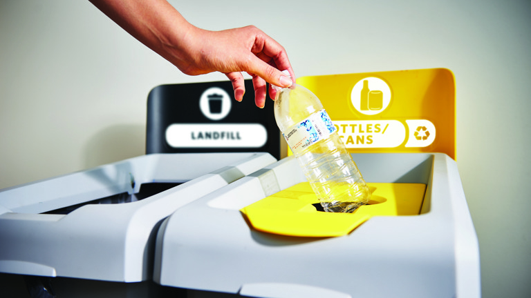 Hand placing plastic bottle into yellow recycling bin labeled ""Bottles/Cans"" next to a black landfill bin."