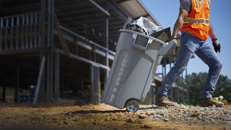 Construction worker in a neon vest moving a large gray garbage bin full of debris on a dusty construction site.