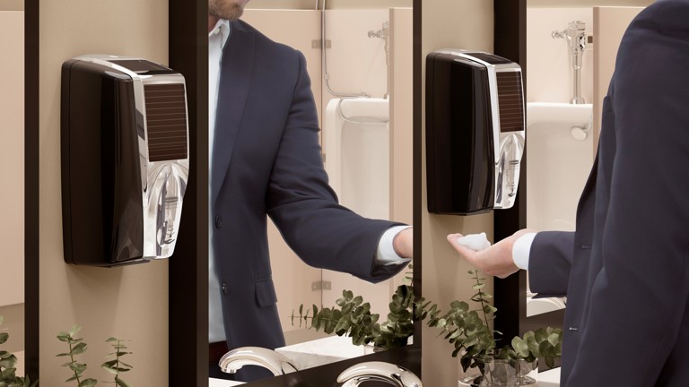 Person in a suit using a modern automatic soap dispenser mounted on a mirror in a restroom with plants on the counter.