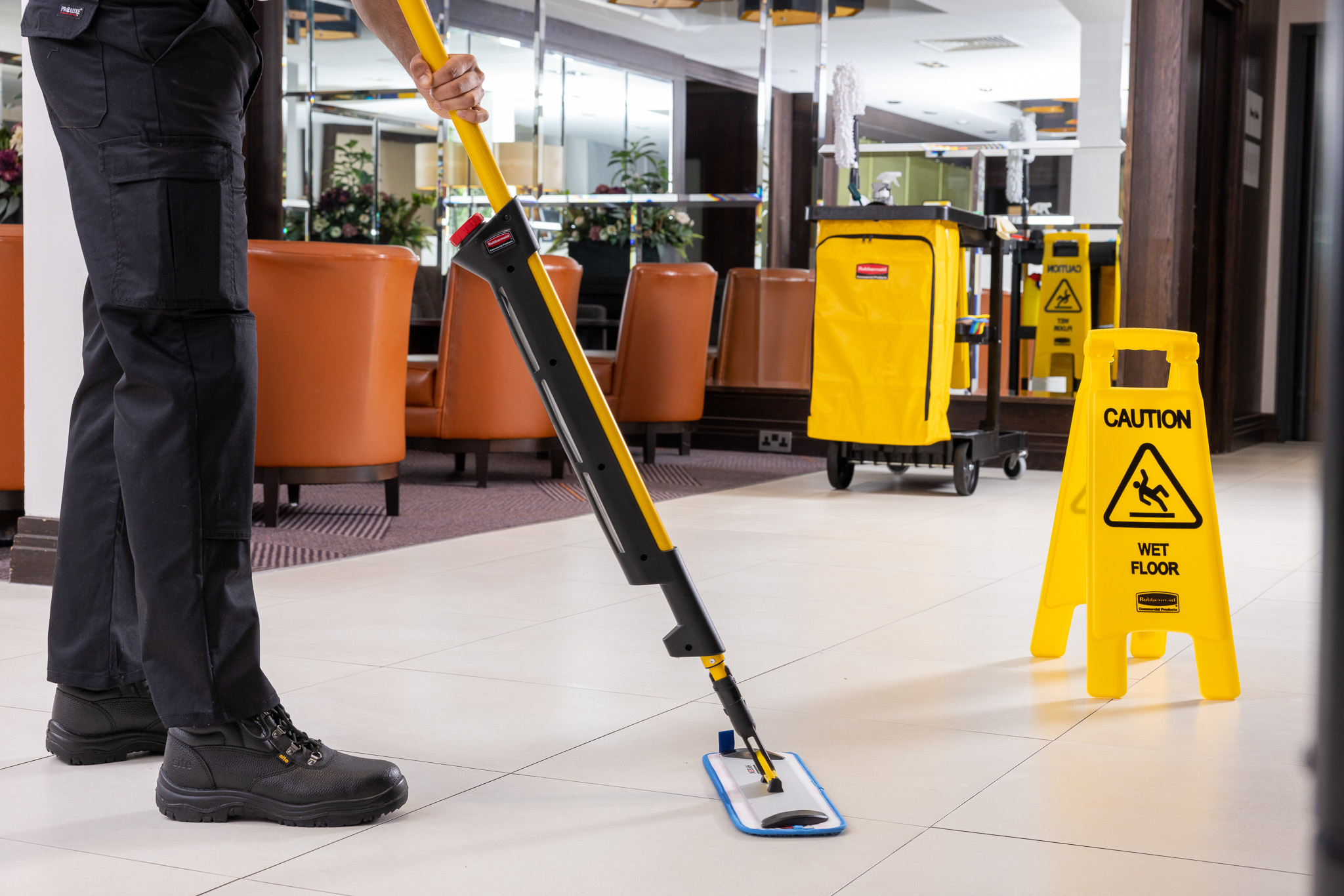 Janitor mopping floor in lobby with caution wet floor sign and cleaning cart nearby.