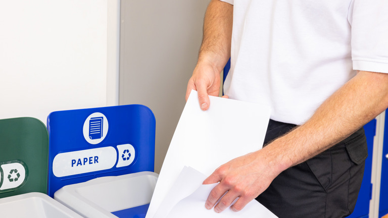 Person placing white paper into a blue recycling bin labeled for paper.