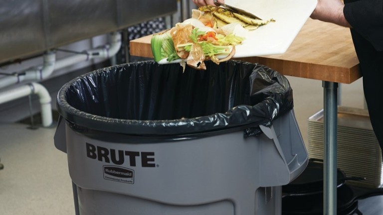 Food waste being disposed into a large gray garbage bin labeled ""BRUTE"" in a kitchen setting."