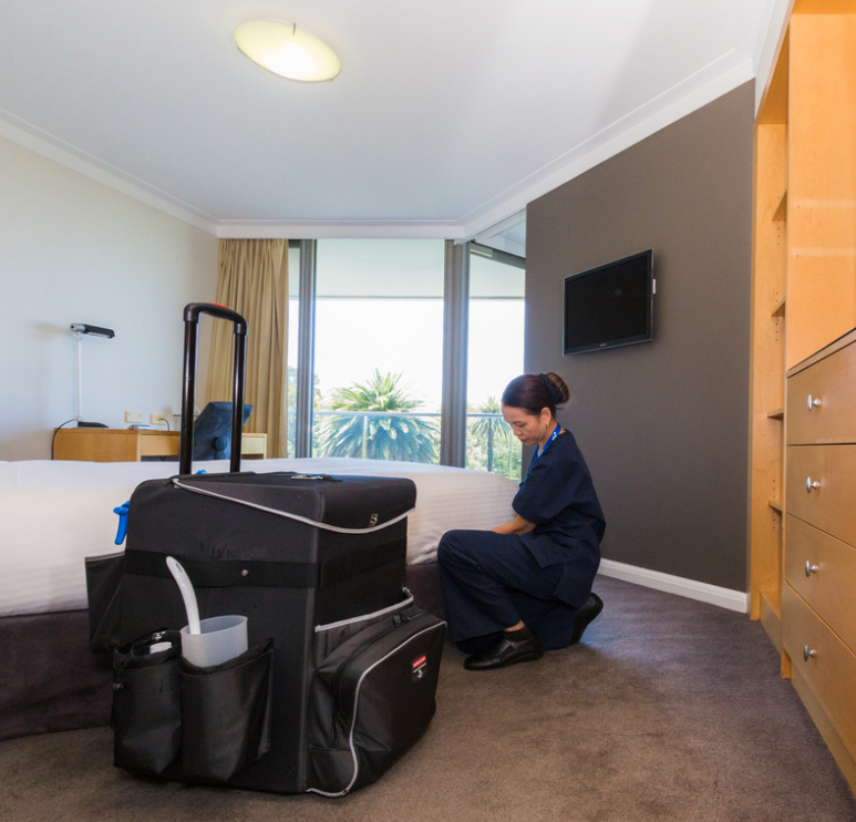 Hotel room with housekeeping staff cleaning near bed; cleaning cart in foreground