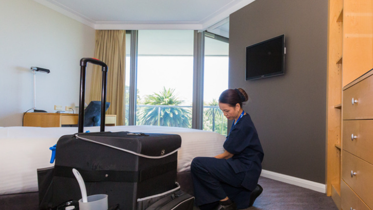 Hotel room with housekeeping staff cleaning near bed; cleaning cart in foreground