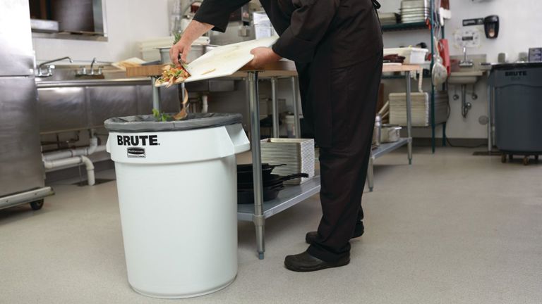 Kitchen worker discarding food scraps into a Brute trash can in a commercial kitchen setting.