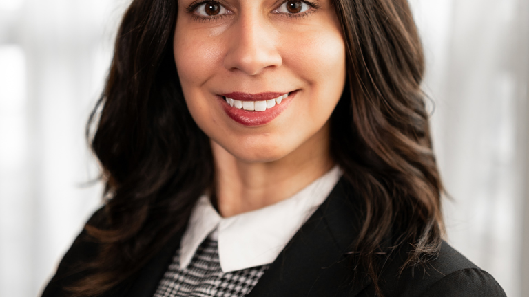 Smiling woman with long brown hair wearing a black blazer and checkered blouse against a blurred background.