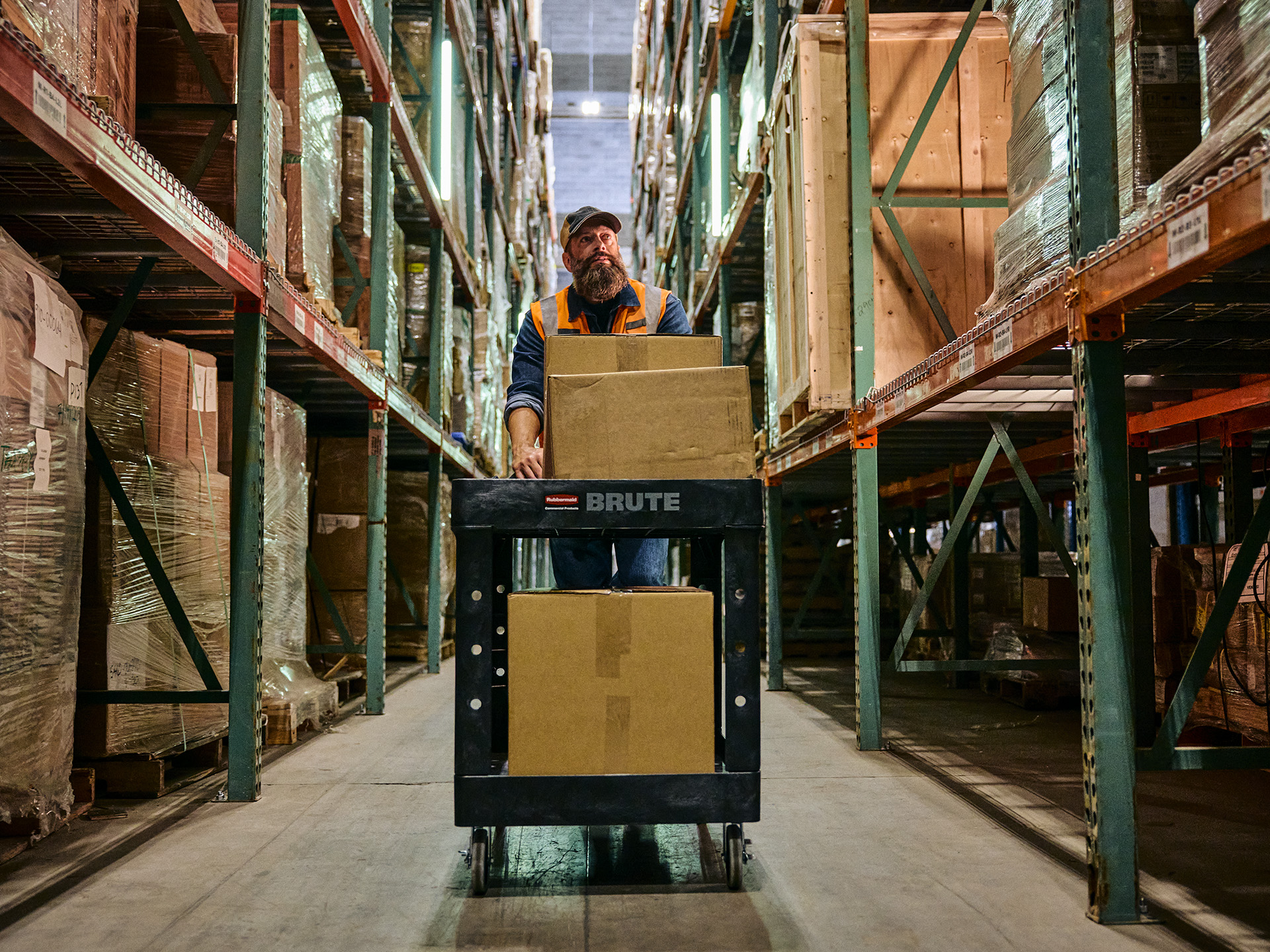 Warehouse worker transporting large boxes on a cart through aisles of stacked shelves.