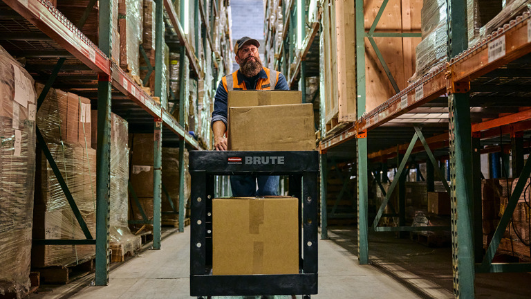 Warehouse worker transporting large boxes on a cart through aisles of stacked shelves.