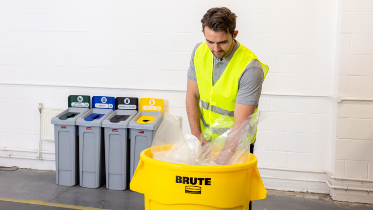 Worker in reflective vest sorts plastic waste into a large yellow bin labeled ""Brute"" with a row of recycling stations in the background."