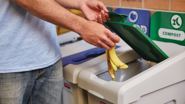 Person composting banana peel in green-lid bin next to recycling containers